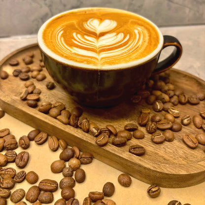 A latte with a leaf poured in the foam, on a wooden tray scattered with roasted coffee beans