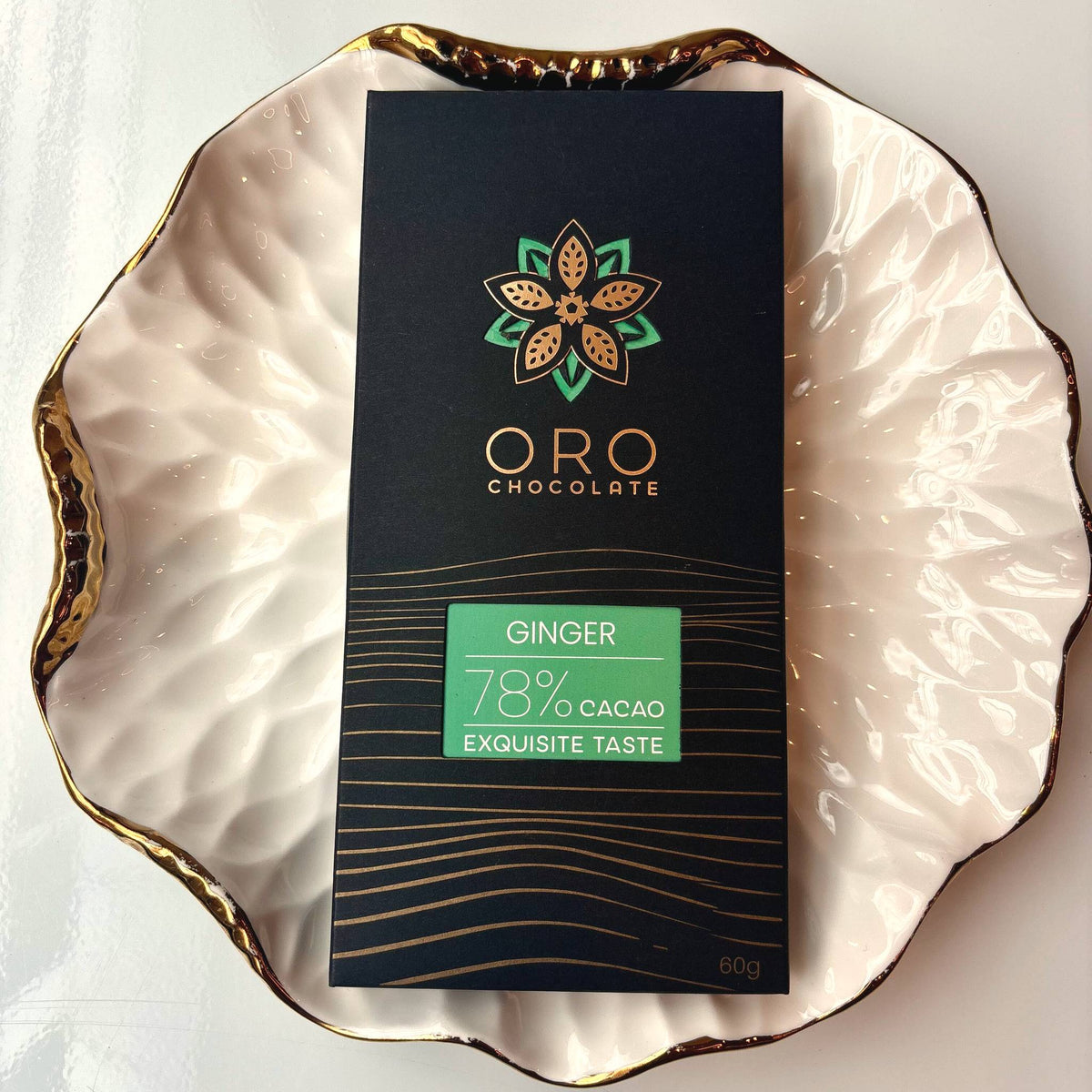 Oro Ginger 78% cacao bar in black packaging on a gold-rimmed white porcelain plate, seen from above