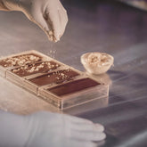 Gloved hands sprinkling toppings onto chocolate bars in a clear mold