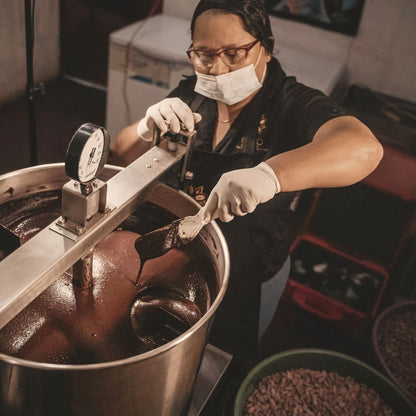 A chocolate maker checking liquid chocolate in a refining tank, cacao nibs in a bowl beside it, Nicaragua
