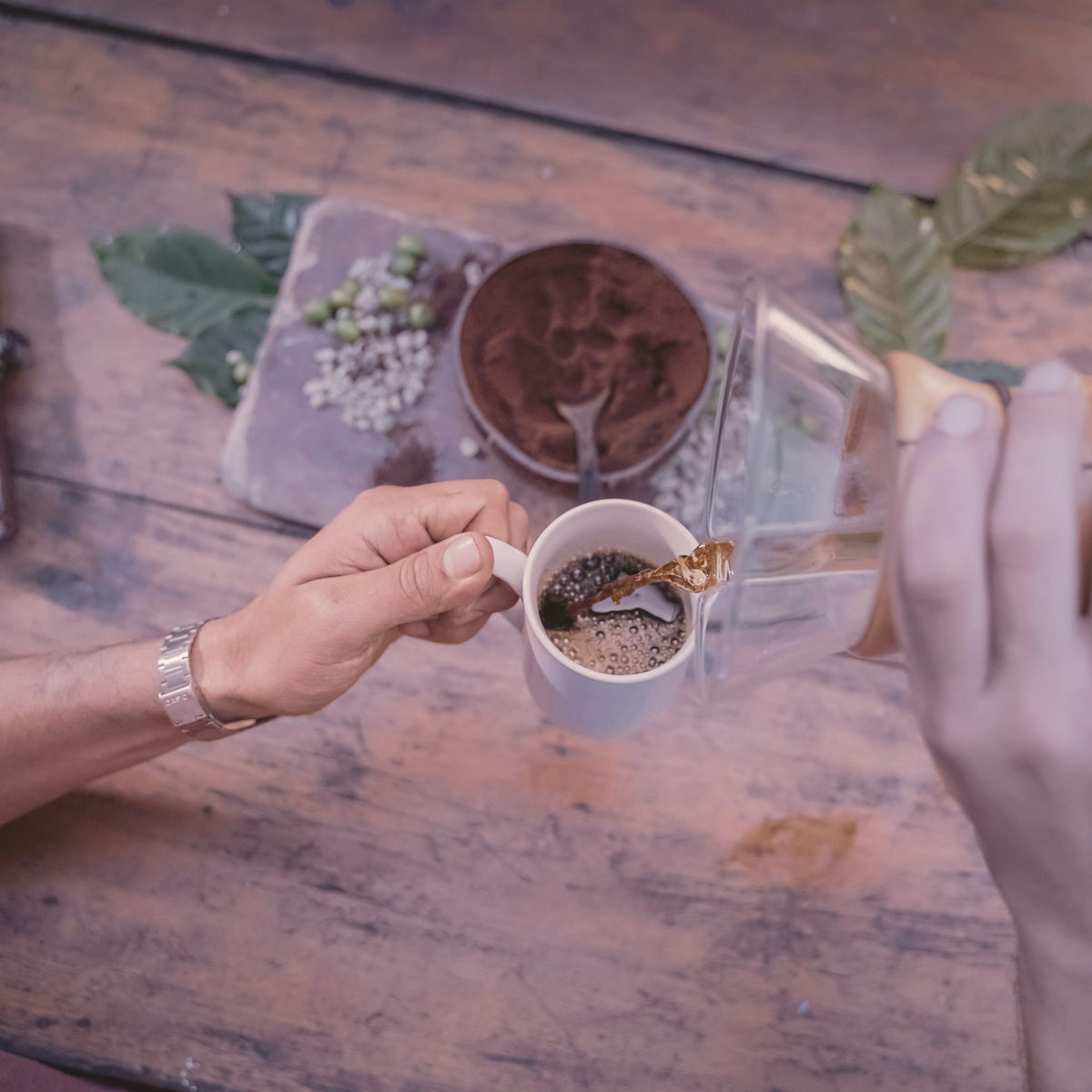 Hands pouring coffee into a small cup on a wooden table, seen from above