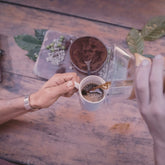Hands pouring coffee into a small cup on a wooden table, seen from above