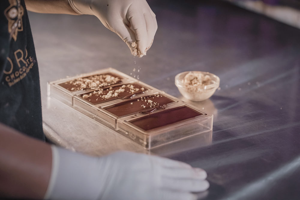 Gloved hands sprinkling toppings onto chocolate bars in a clear mold.