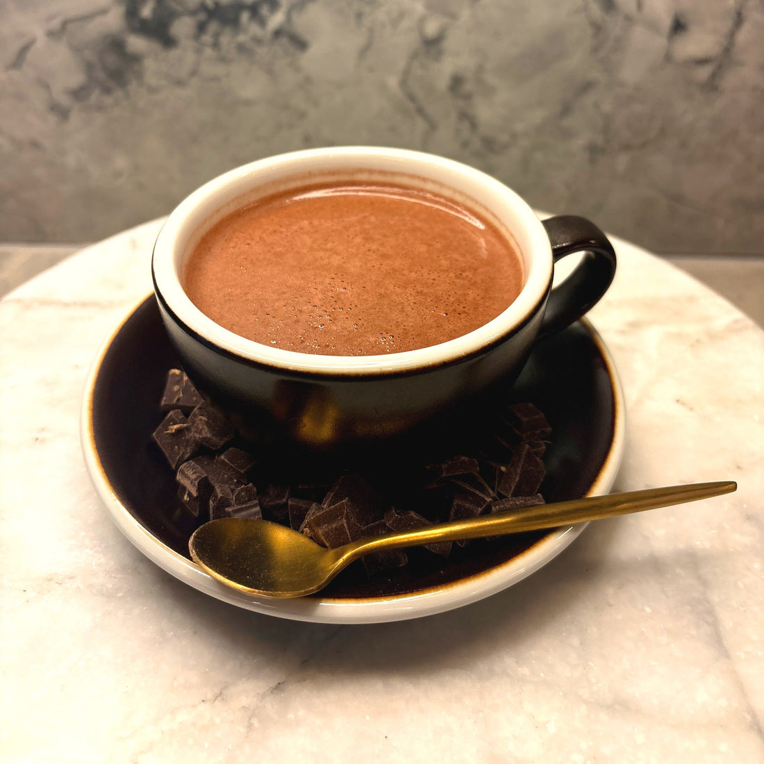 A cup of Oro drinking chocolate on a saucer scattered with chocolate pieces and a gold spoon