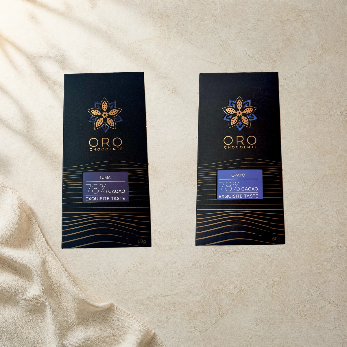 Editorial arrangement of Oro Tuma and Opayo 78% cacao bars in original black and gold packaging, 60g each, on cream stone.