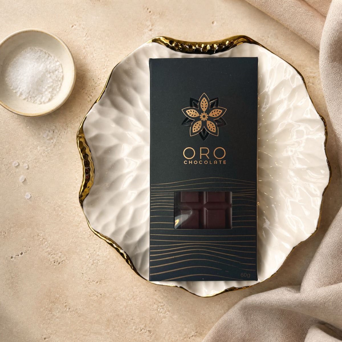 Oro Sea Salt 78% cacao bar in its original black windowed pack on a porcelain plate, with flaky sea salt, linen and cream stone