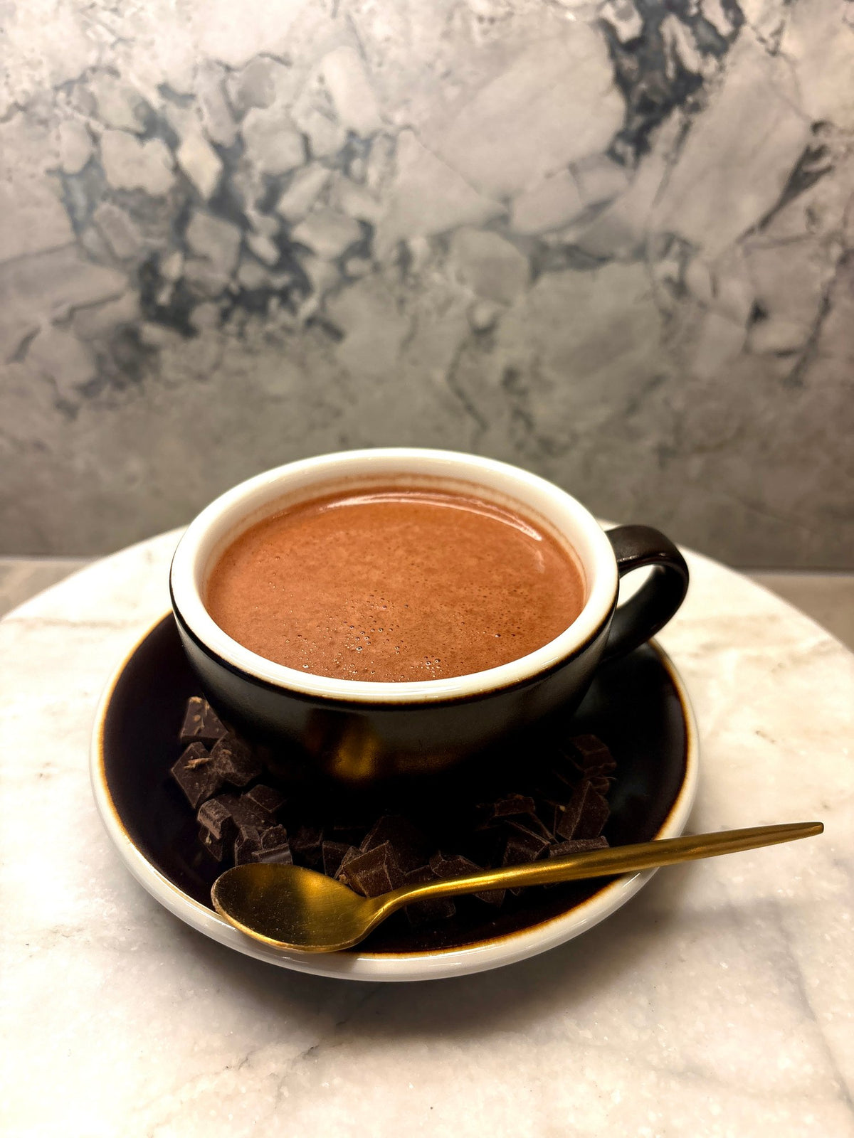 A cup of Oro drinking chocolate on a saucer scattered with chocolate pieces and a gold spoon