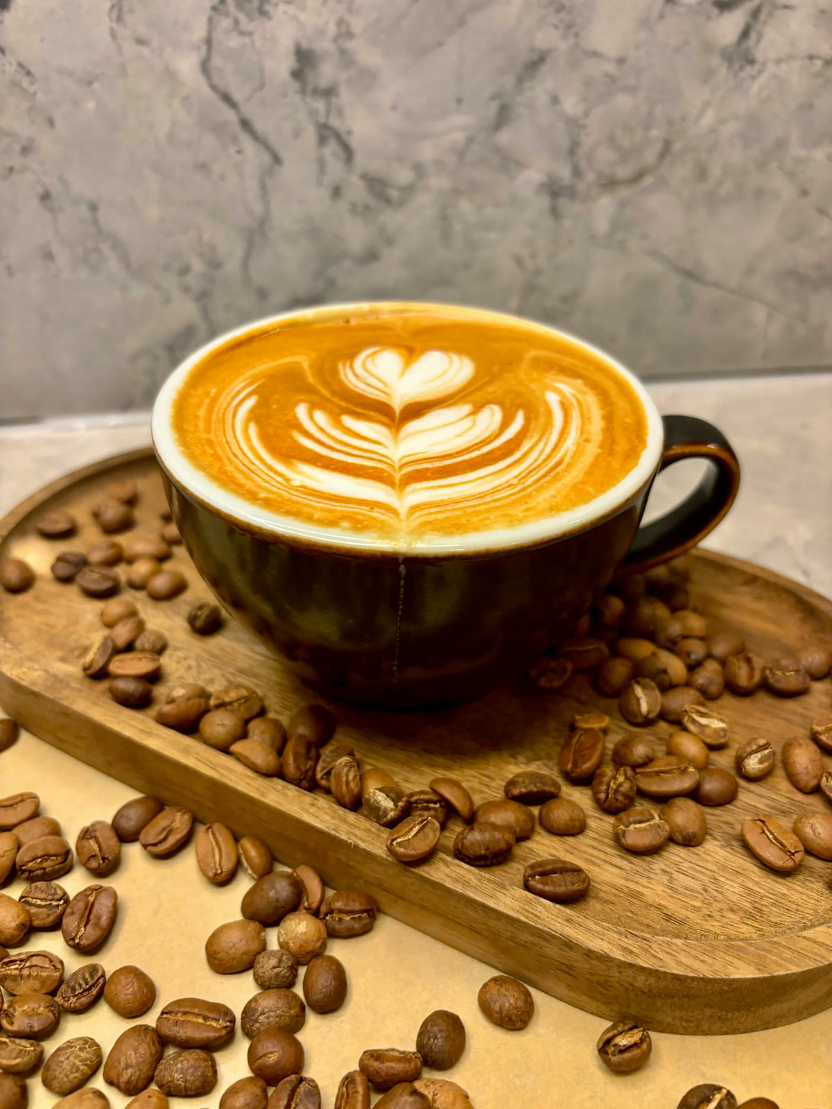 A latte with a leaf poured in the foam, on a wooden tray scattered with roasted coffee beans