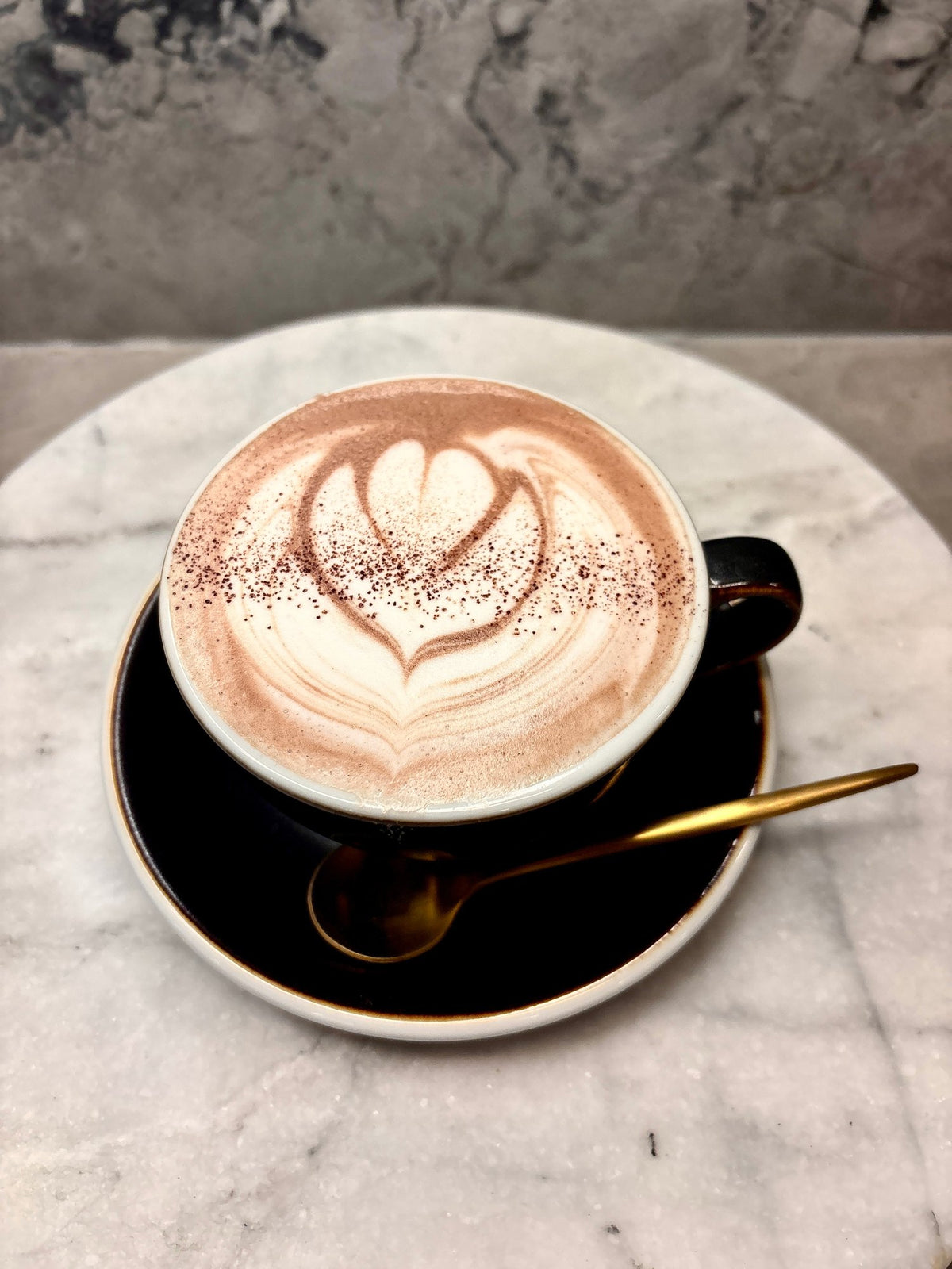 Oro drinking chocolate seen from above, a heart poured in the foam, on a black saucer with a gold spoon