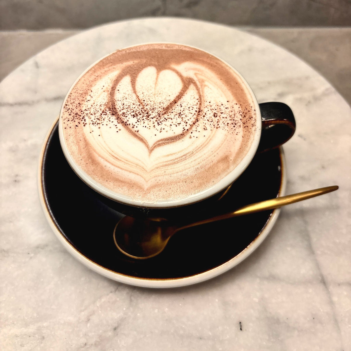 Oro drinking chocolate seen from above, a heart poured in the foam, on a black saucer with a gold spoon